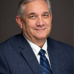 A man looks into a camera. He is wearing a blue suit and tie.